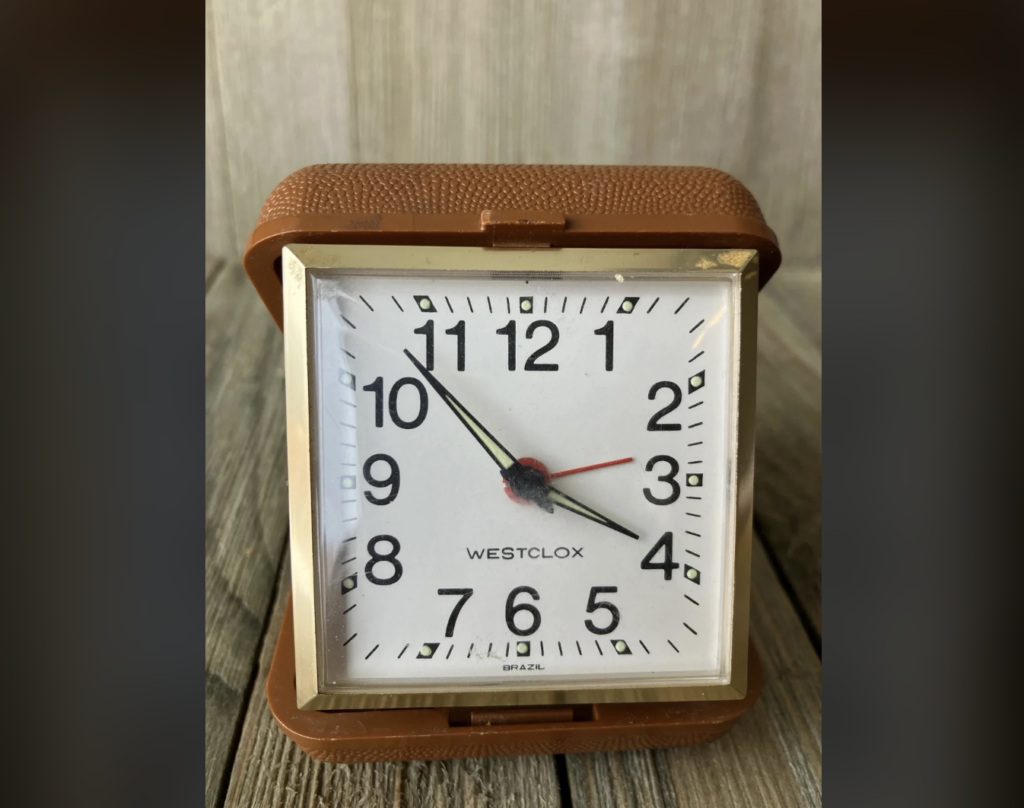 A brown, vintage Westclox alarm clock with a white face and black Arabic numerals. The clock has black hour and minute hands, a thin red second hand, and is set to approximately 7:17. The clock is placed on a wooden surface.