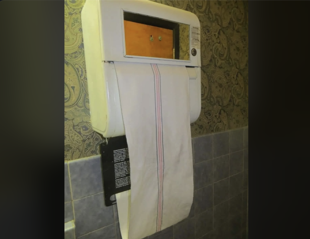 A wall-mounted automatic hand dryer with a continuous piece of cloth towel. The cloth hangs down for drying hands. The background features beige patterned wallpaper and gray tiled walls.