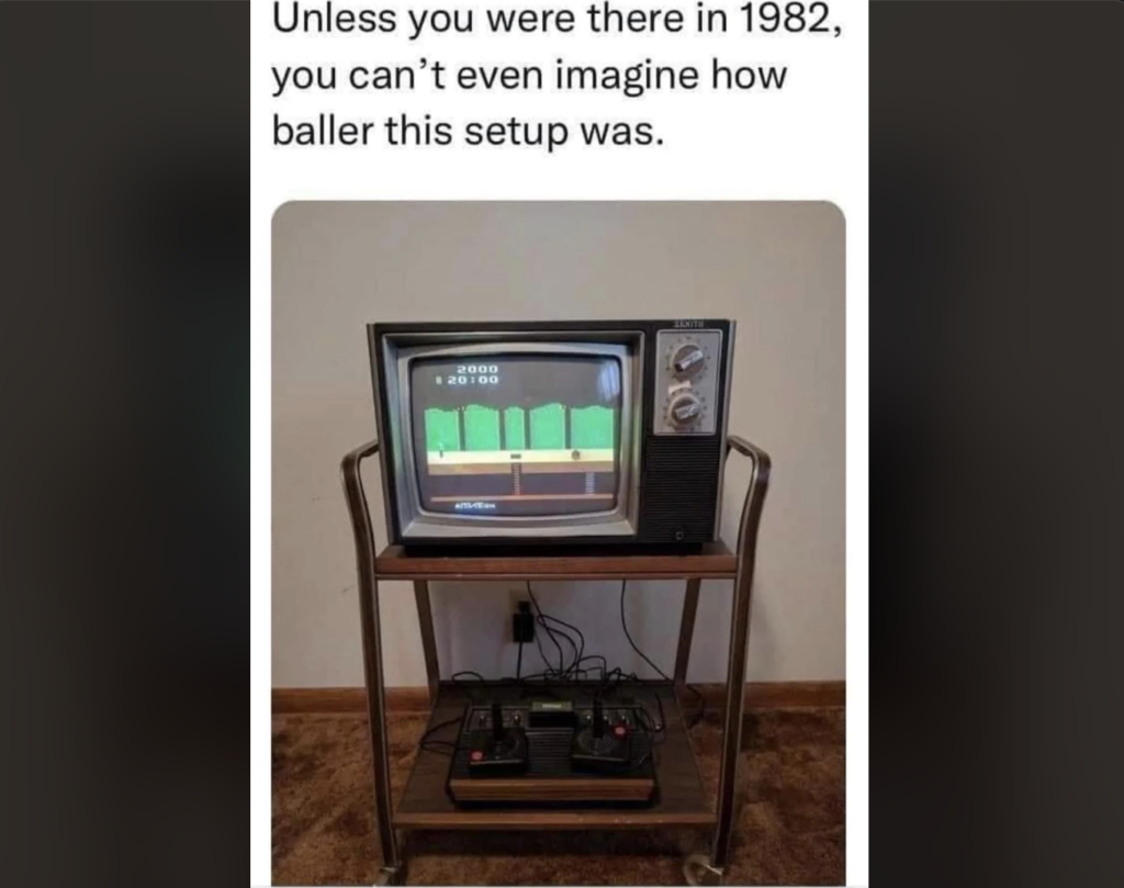 A vintage television from the 1980s sits on a metal and wood TV stand. On the screen is a retro-style video game. Below the TV, there is an old game console with two joystick controllers attached. Text above reads, "Unless you were there in 1982, you can't even imagine how baller this setup was.