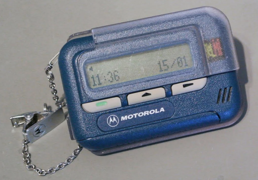 A blue Motorola pager with a grey clip-on chain is displayed. The screen shows the time 11:36, and below it, another time which appears to be 15:01. The pager has buttons below the screen and the brand logo on the front.