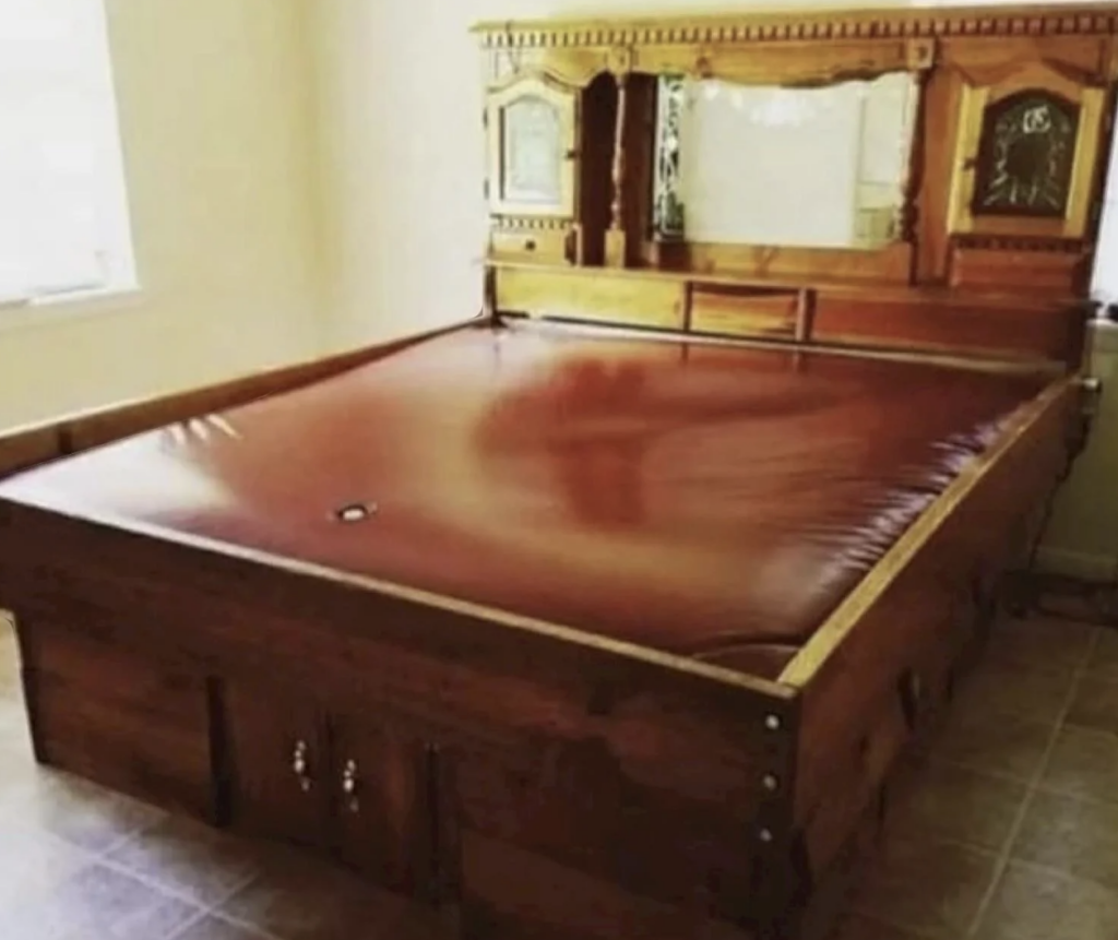 A wooden-framed waterbed with intricate carvings on the headboard, featuring a built-in mirror and small storage compartments. The room has tiled flooring and a window allowing natural light to enter. The waterbed appears to have a smooth, red-tinted surface.