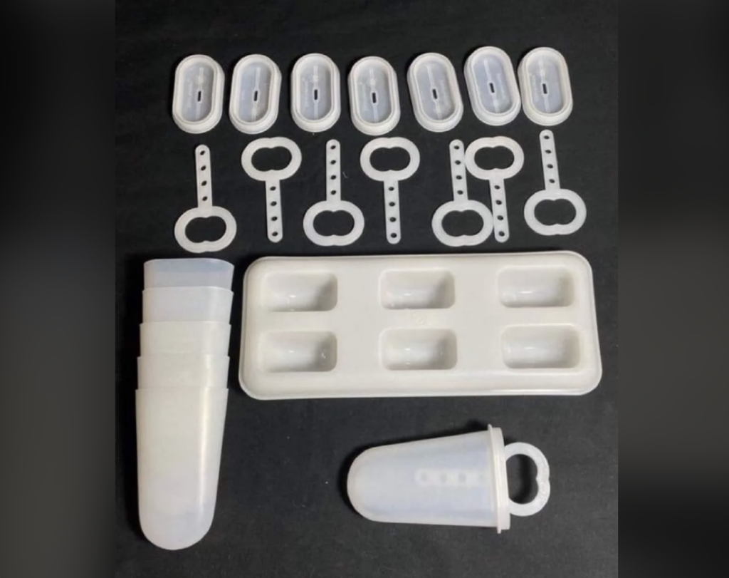 A white popsicle mold set displayed on a black surface. The set includes a tray for six popsicles, four molds with pointed tips, and six handles with stoppers. The molds and handles are arranged neatly above and beside the tray.