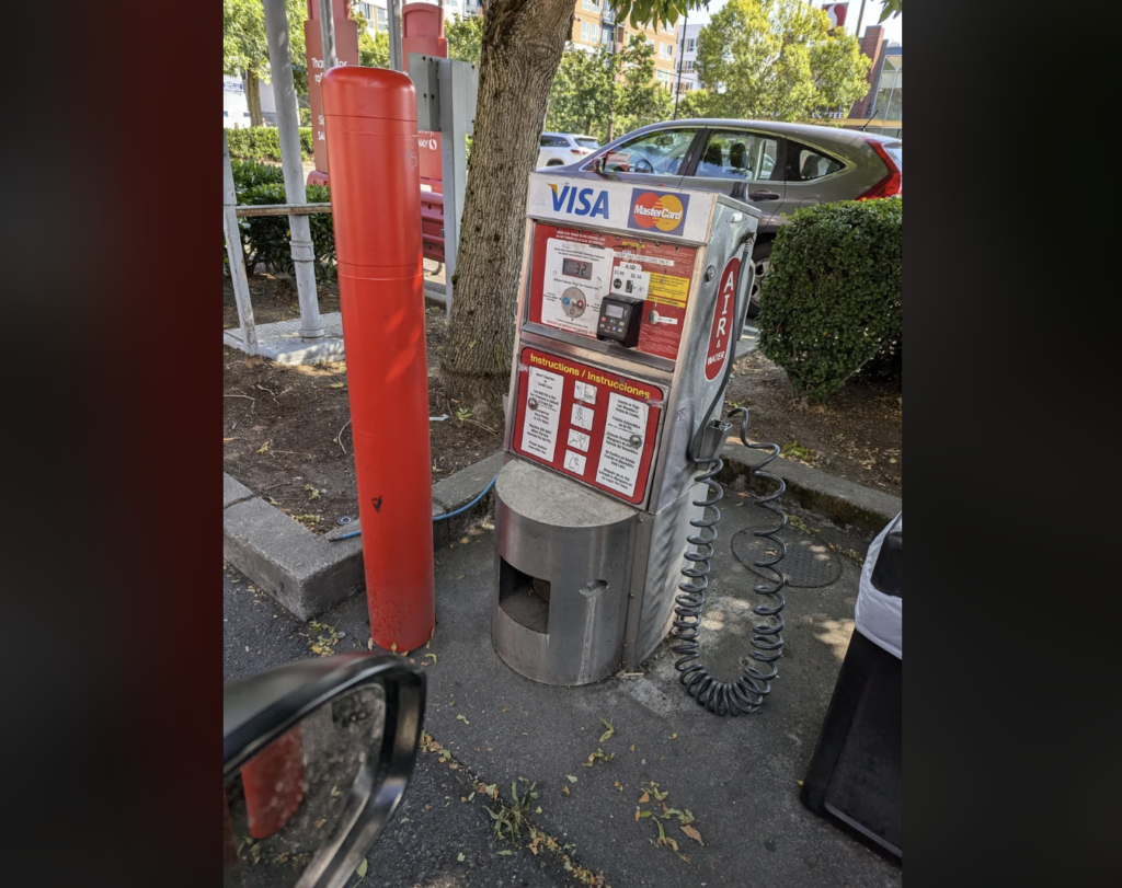 A public air pump station for vehicle tires located in a shaded area beside a tree. The station features payment options including Visa and MasterCard. It is equipped with a hose and gauge for inflating tires. A red cylindrical bollard is nearby.