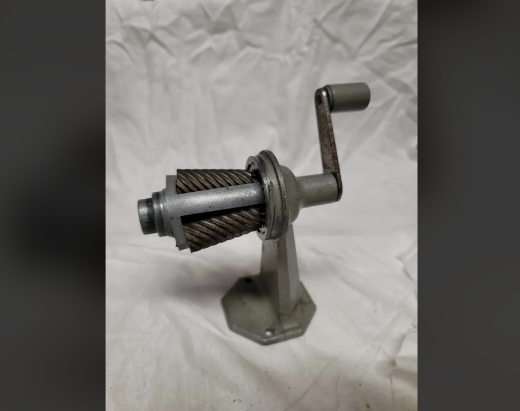 A vintage hand-crank tool with a cylindrical brush attachment, mounted on a base. The tool appears to be made of metal with a rustic finish, and is set against a wrinkled white fabric background.