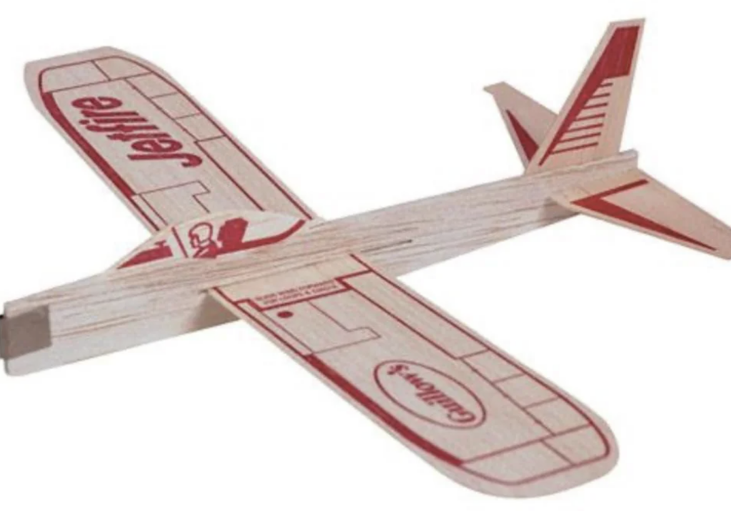 A small model glider made of lightweight wood with red printed designs, including the word "Jetfire" on the wings. The glider has a long fuselage, wide wings, and a tail section with vertical and horizontal stabilizers.