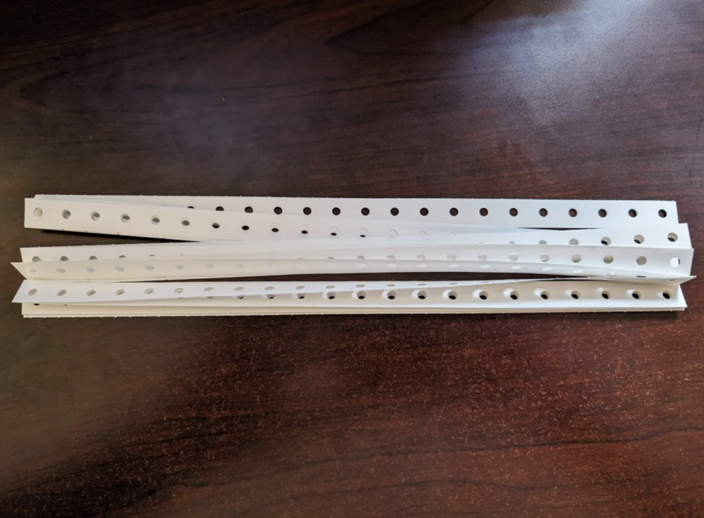 A stack of white, perforated plastic strips lies on a dark wooden surface. The strips are aligned with several small, evenly spaced holes running along their length.