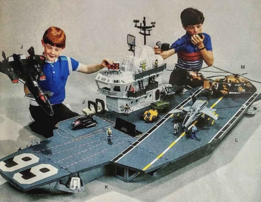 Two boys play with a large aircraft carrier toy set, complete with fighter jets, a control tower, and various miniature vehicles and action figures. One boy is wearing a red shirt and is holding a black jet, while the other boy, in a blue shirt, is playing with the control tower.