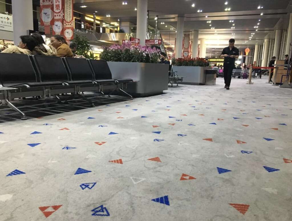 A spacious airport terminal features geometric-patterned carpet. Travelers sit on black chairs near a planter as someone walks by, their footsteps echoing in the modern, airy space. Soft lighting and signage hang from the ceiling, enhancing the bustling yet calm airport atmosphere.