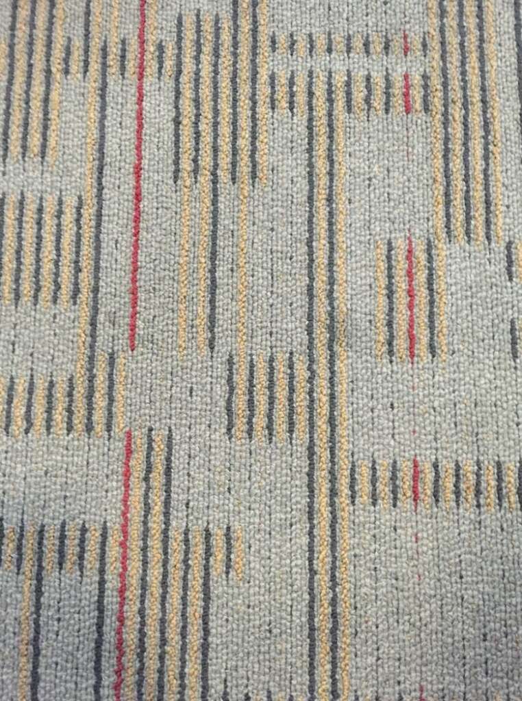 Carpet with a geometric pattern featuring vertical and horizontal lines in shades of beige, red, and gray on a light gray background, reminiscent of the sleek designs often found in airport terminals.