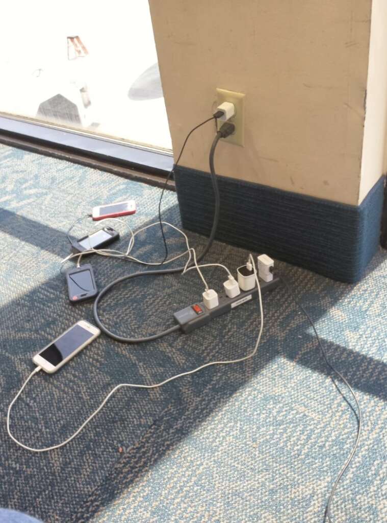 Several smartphones and chargers are plugged into a power strip connected to a wall outlet, mimicking an airport charging station, as they rest on a blue carpet next to a window.