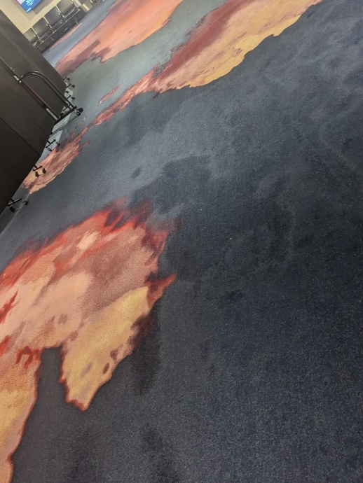 A carpeted floor with an abstract design featuring orange, red, and yellow patterns on a dark background evokes the vibrant energy of an airport terminal. A railing or barrier is partially visible on the left side, hinting at the bustling environment beyond.