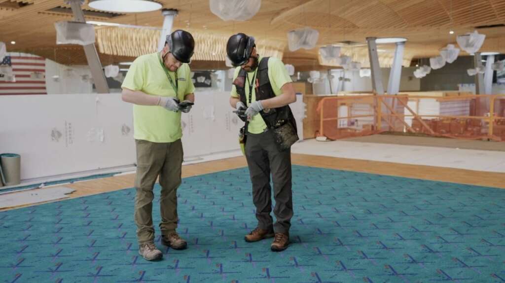 Two construction workers in safety gear are meticulously examining papers or electronic devices on a patterned carpet. The indoor scene resembles an active airport terminal under renovation, with construction materials and equipment clearly visible in the background.