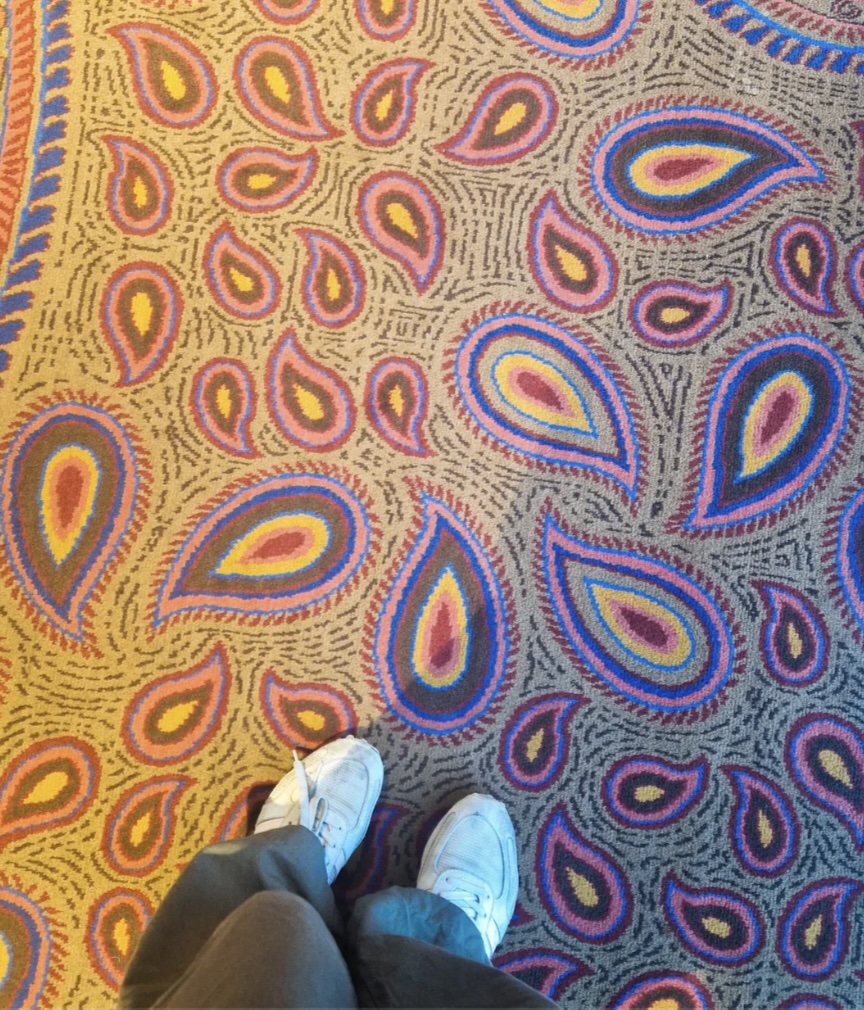 The image shows a pair of feet in white sneakers standing on a brightly colored, paisley-patterned airport carpet, featuring vibrant blue, pink, and orange hues.