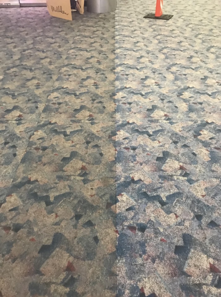 Blue and gray patterned carpet at the airport shows a noticeable difference in color and texture on each side, indicating a cleaner or newer section. A traffic cone is visible in the background.