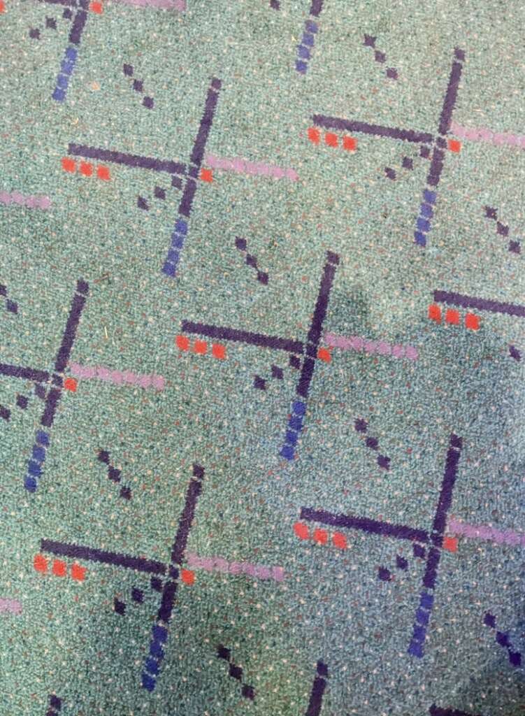 A close-up of a patterned carpet in an airport showcases a teal background with intersecting lines and dots in navy blue, red, purple, and black. The design creates a geometric grid across the surface, capturing the dynamic energy of travel hubs.