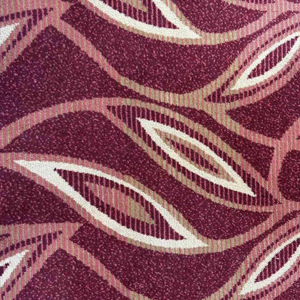 A close-up of a carpet with an abstract pattern, reminiscent of those found in airport lounges. The design features wavy lines and oval shapes in shades of maroon, beige, and pink, creating an intricate and textured appearance.