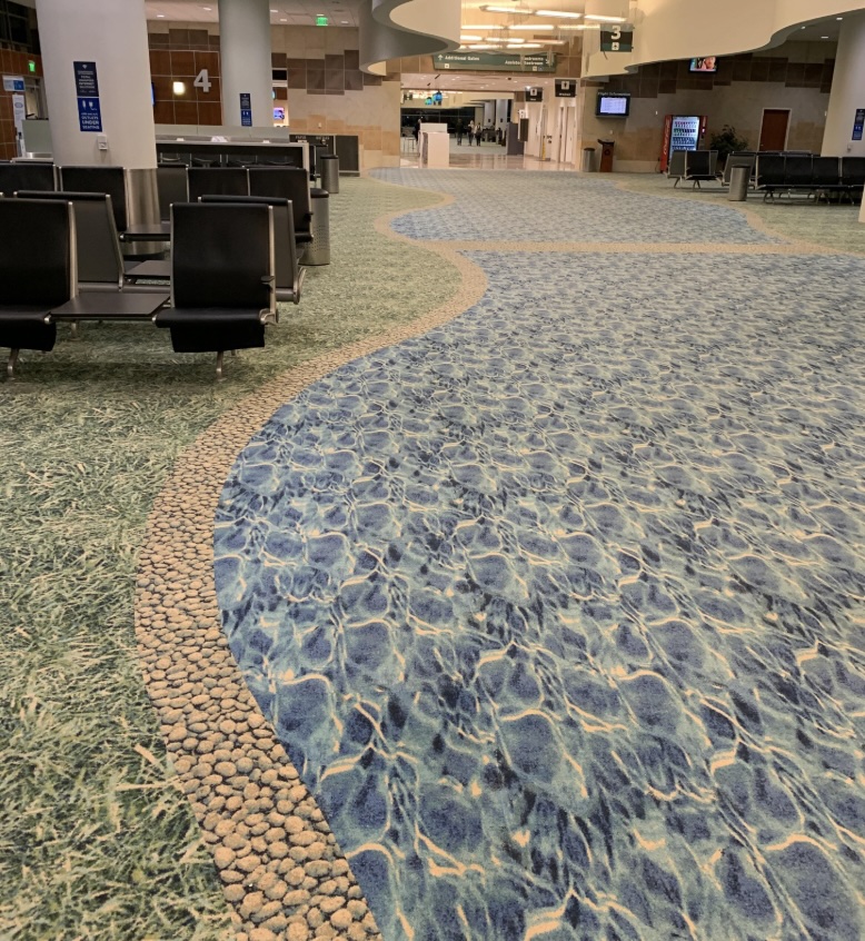 The airport terminal boasts a unique carpet design resembling water and pebbles. Empty black chairs line the left side, while signs with flight information hang from the ceiling. This brightly lit and spacious area enhances the overall airport experience.