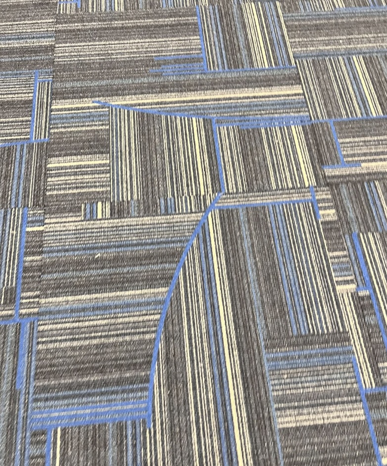 A close-up of a carpet with a geometric pattern reminiscent of airport terminal designs. The carpet showcases gray, blue, and cream stripes in varying widths, arranged in squares and rectangles. A curved blue line intersects a few of the shapes, adding dynamic flair.