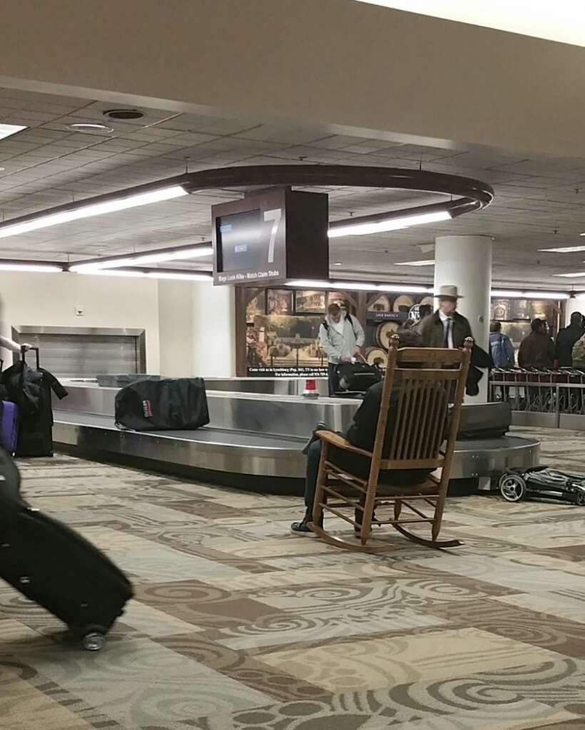 A person in a rocking chair closely observes the luggage carousel at the airport baggage claim, while another stands nearby with a suitcase. The well-lit area features a display screen indicating carousel number 7.