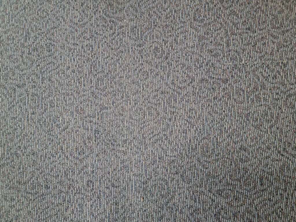 Close-up of a gray carpet reminiscent of those found in airport terminals, featuring a subtle, textured pattern. The surface shows slight variations in color, with hints of blue and beige. The fibers create a uniform and structured appearance.