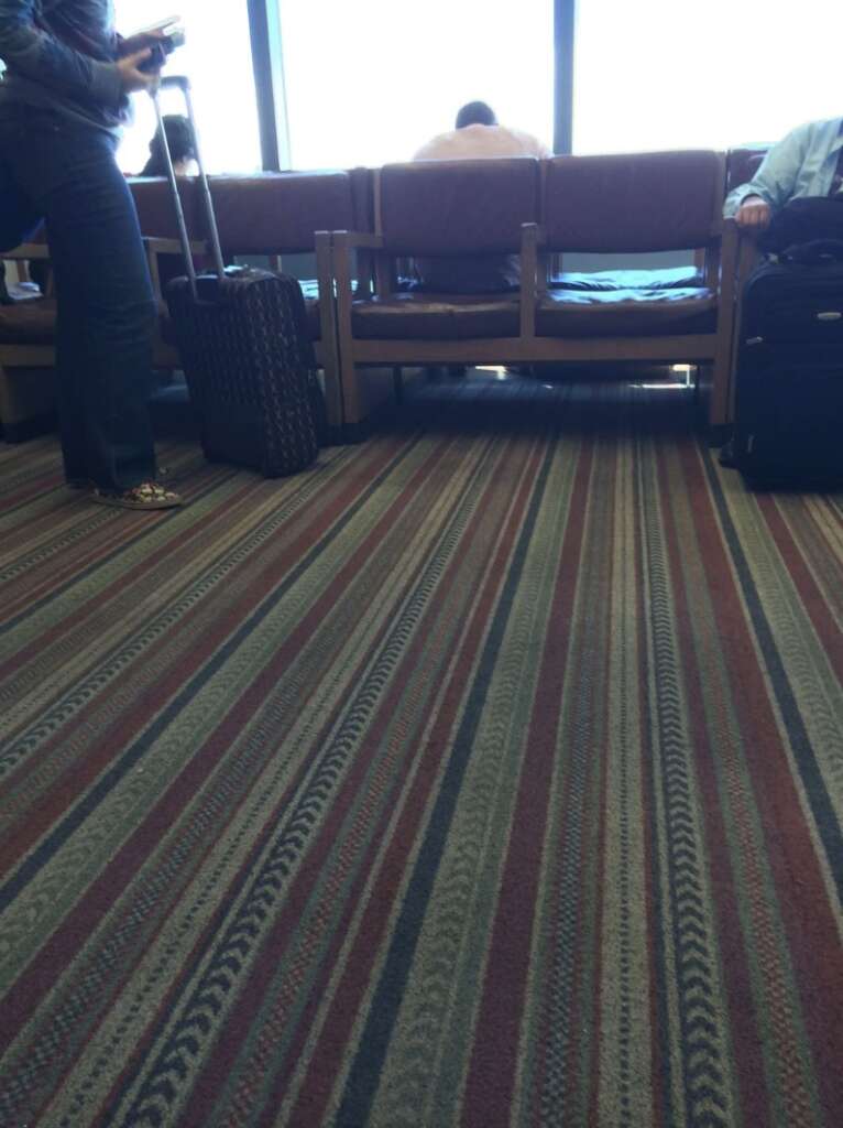 The airport's waiting area features patterned carpeting, where travelers, with luggage in tow, are seated and standing. They gaze through large windows, one person's back turned as they watch planes taxi on the runway.