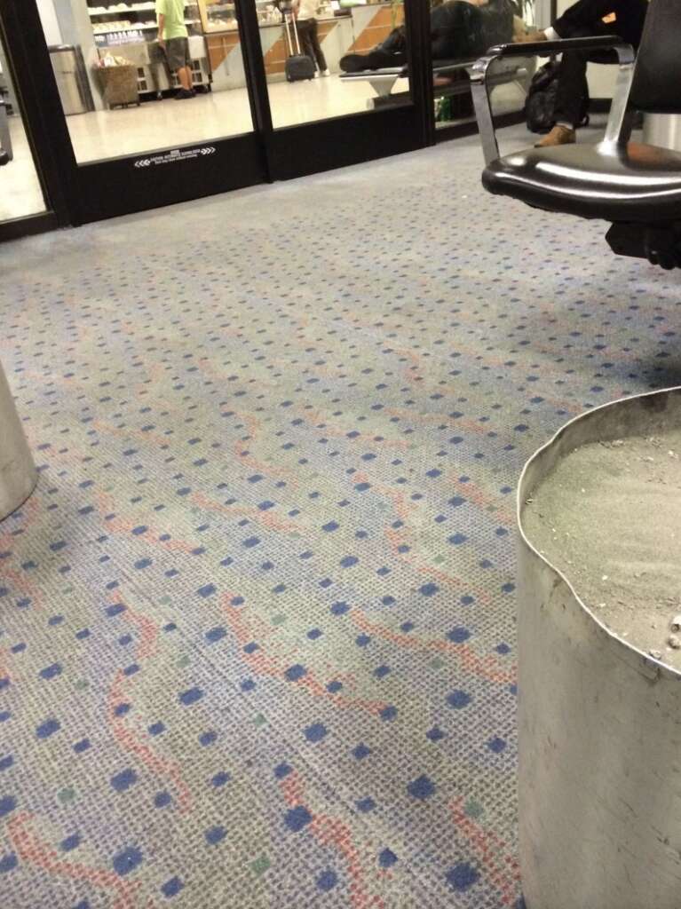 The airport waiting area features a patterned carpet, an empty metal ashtray, and dark chairs. Glass doors in the background reveal a vending area. A few people can be glimpsed through the shimmering glass.