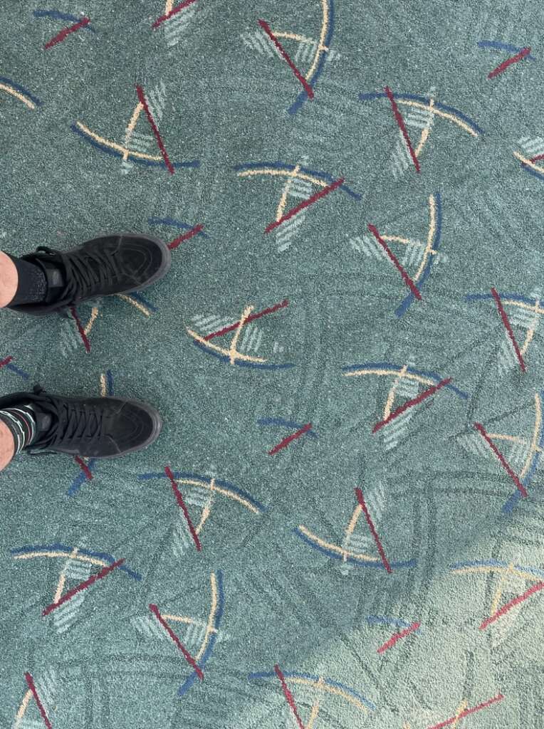 A person wearing black sneakers stands on a patterned carpet reminiscent of those found in an airport, featuring red, blue, and white geometric designs set against a teal background.