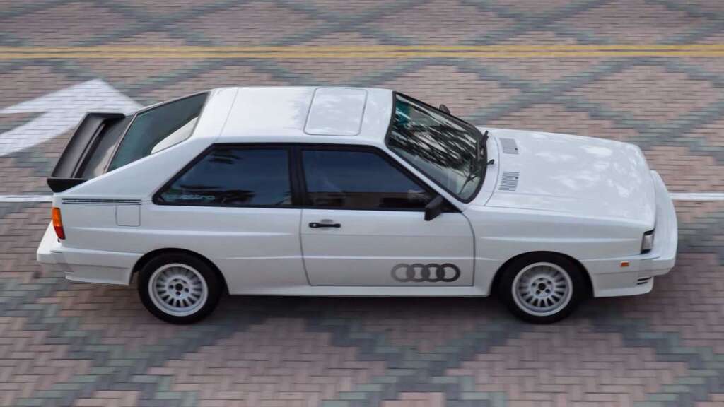 A white sports car with a large rear spoiler and distinctive side decals, reminiscent of the coolest cars from the 1980s, is parked on a patterned brick surface. Its boxy shape evokes a nostalgic era of automotive design, further highlighted by a white arrow pointing left.