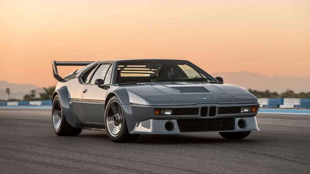 A sleek, silver sports car reminiscent of the coolest cars from the 1980s is captured on a racetrack at sunset. The sky is a soft orange, and the car's aerodynamic design highlights its performance focus. The background shows distant mountains and a blurred track.