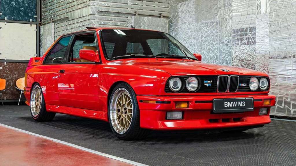 A classic red BMW M3 from the coolest cars of the 1980s is parked indoors, showcasing its sleek design. The car features gold alloy wheels and has a distinctive, sporty appearance. The background includes industrial-style walls and a mix of floor textures.