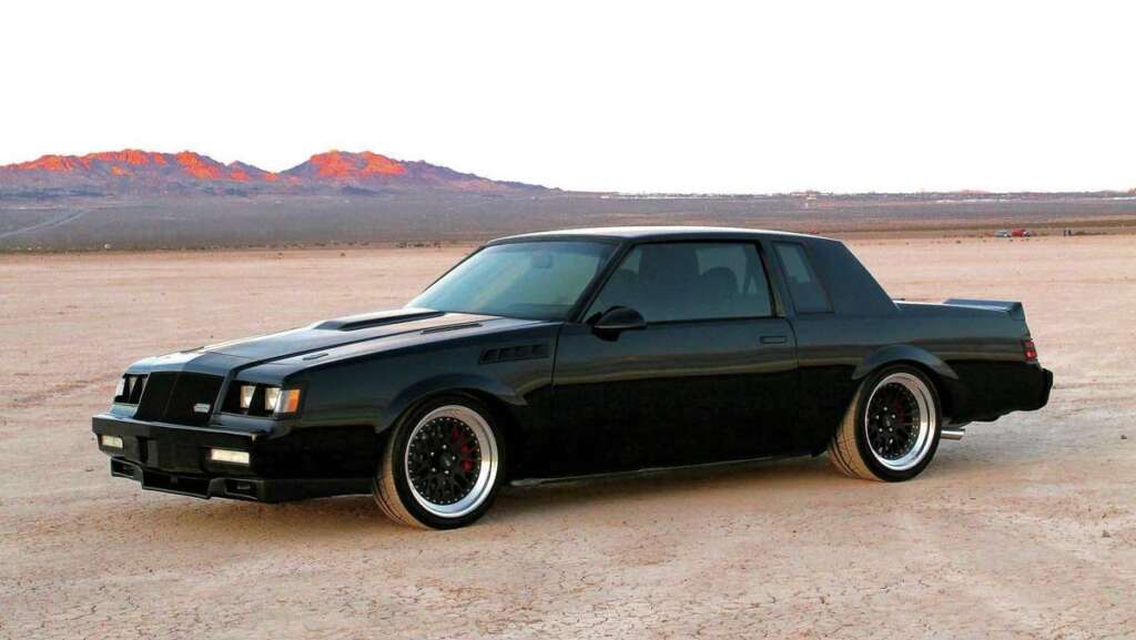 A black, vintage muscle car, one of the coolest cars from the 1980s, is parked on a barren desert landscape with distant mountains under a clear sky. The car boasts a sleek, glossy finish with distinctive wheels and tinted windows.