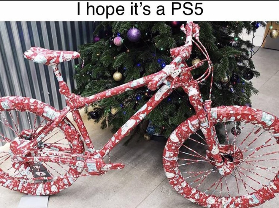 A bicycle completely wrapped in Christmas-themed paper is displayed in front of a decorated Christmas tree. Above the bike is the humorous text, "I hope it's a PS5.