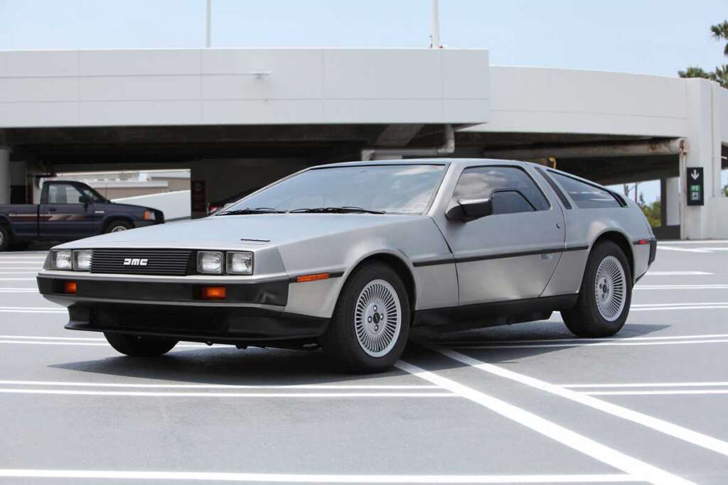 Coolest cars of the 1980s - A silver DeLorean DMC-12, one of the coolest cars from the 1980s, is parked in a rooftop parking lot. The car features iconic gull-wing doors and a distinctive stainless steel body. In the background, there's a partial view of another vehicle and a modern concrete structure.