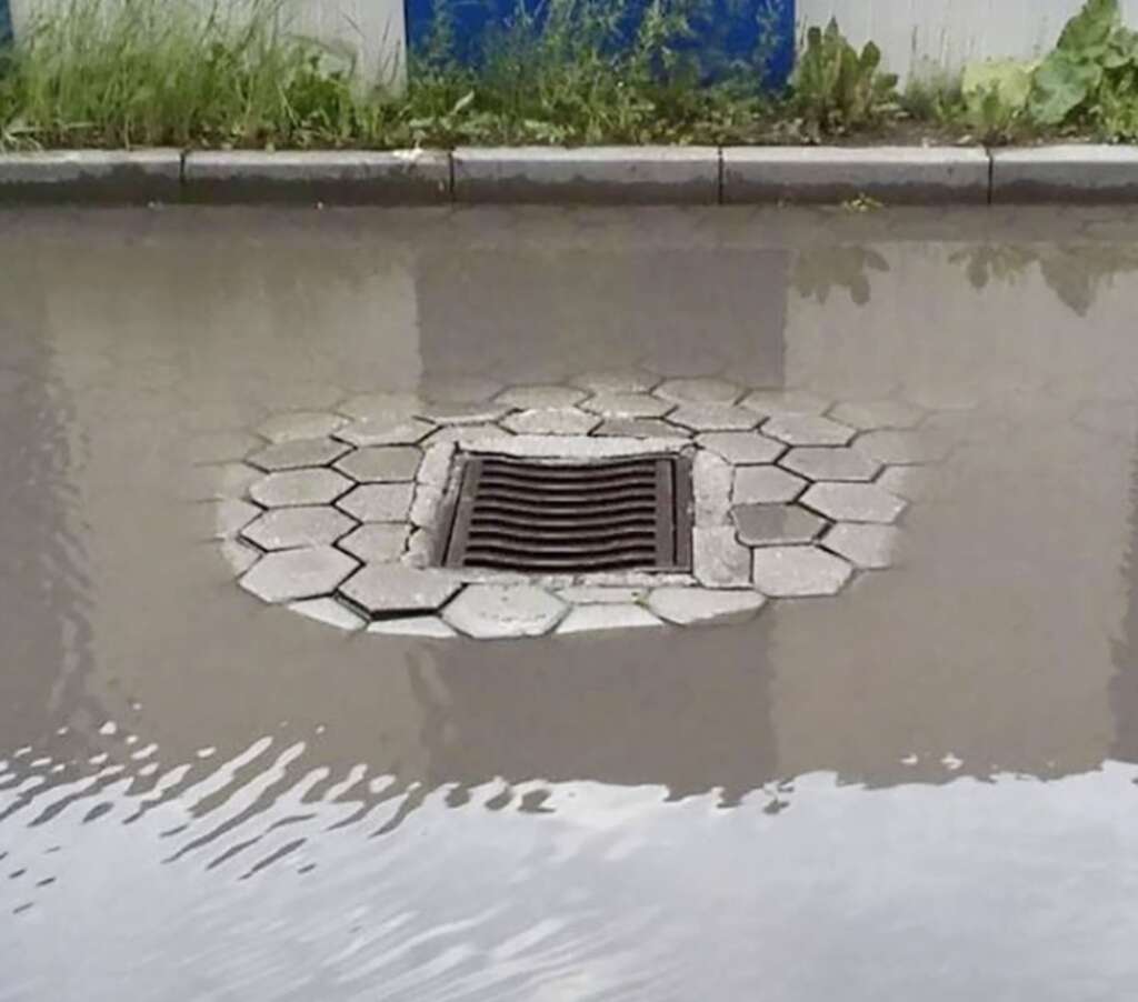 A flooded street with water pooling around a raised storm drain, surrounded by hexagonal pavement tiles. The water reaches the top of the drain, indicating heavy rain or poor drainage. Vegetation is visible along the curb in the background.