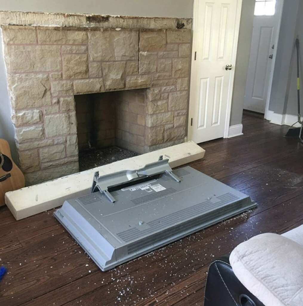 A flat-screen TV lies face down on a wooden floor in front of a stone fireplace. Debris is scattered around. A guitar leans against the wall, and a broom is nearby. The door to another room is slightly open.