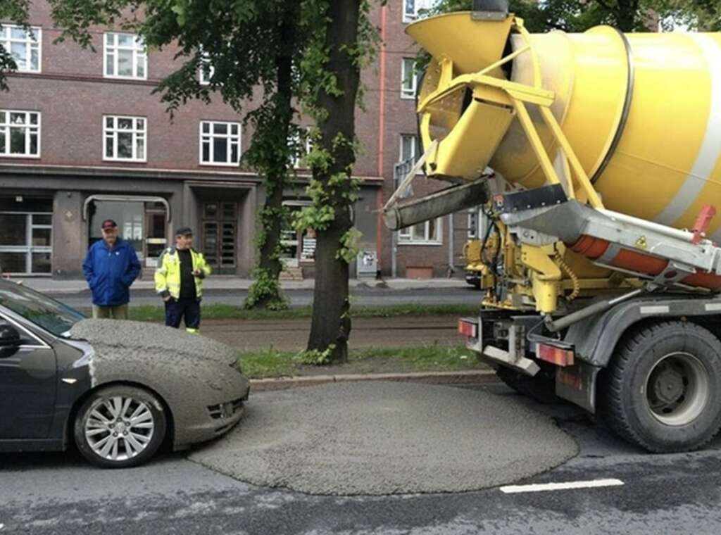 A cement truck accidentally spilled wet cement onto the hood of a parked car on the street. Two workers stand nearby, observing the situation. The background features a building with large windows and trees lining the street.