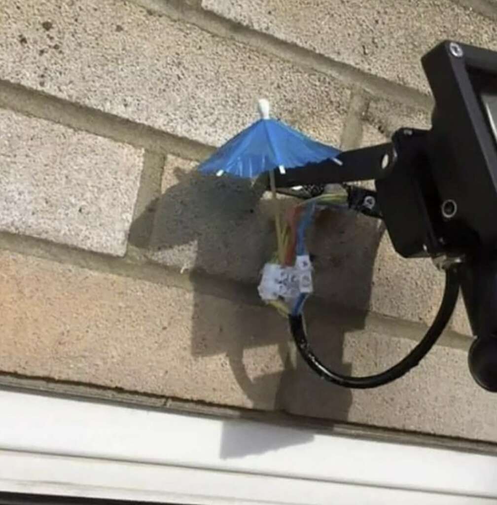 A security camera's exposed wires on a brick wall are covered by a small blue cocktail umbrella to protect them from rain.