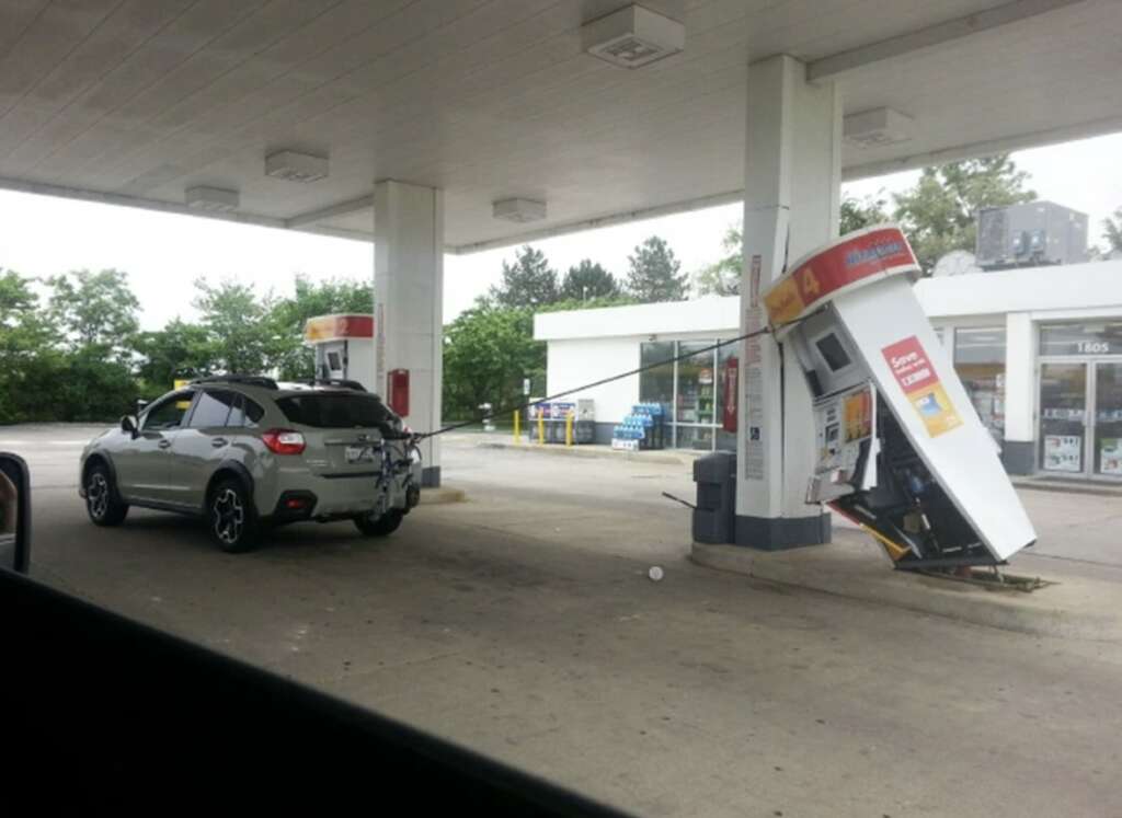 A car is driving away from a gas station with the fuel pump nozzle still attached, pulling over the gas pump. The pump is leaning dramatically, nearly detached from its base. The station is empty, and trees are visible in the background.