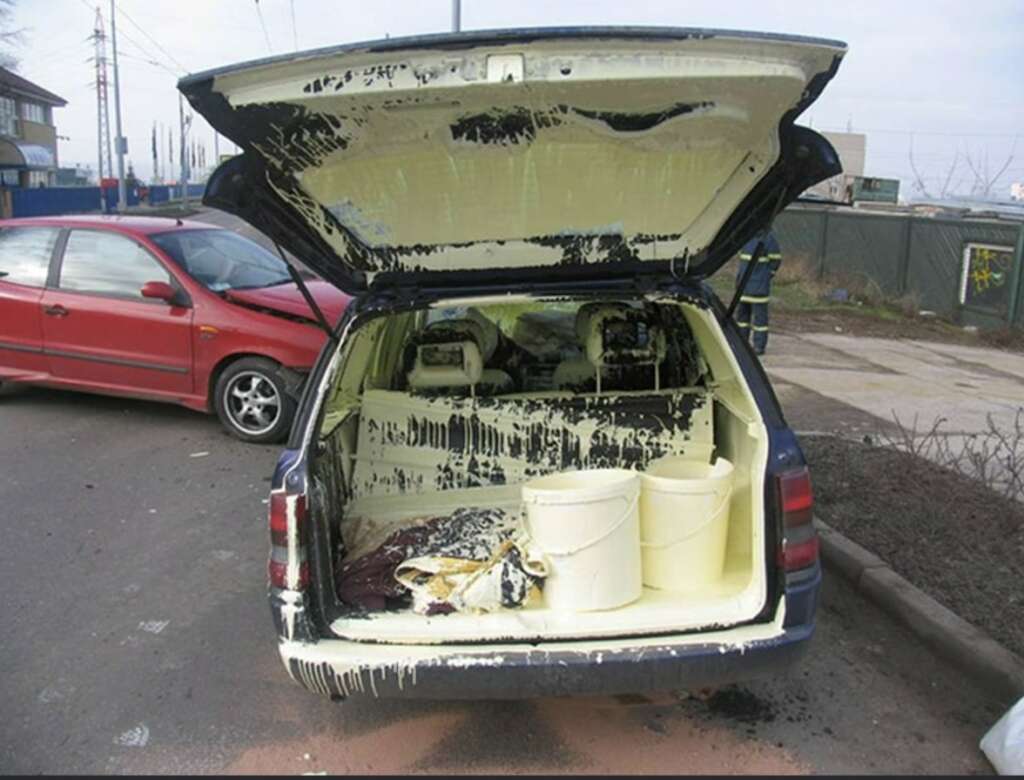 The image shows the open trunk of a car, which is splattered with white paint. Two large buckets, also covered in paint, are inside the trunk. The car is parked on a street near other vehicles.