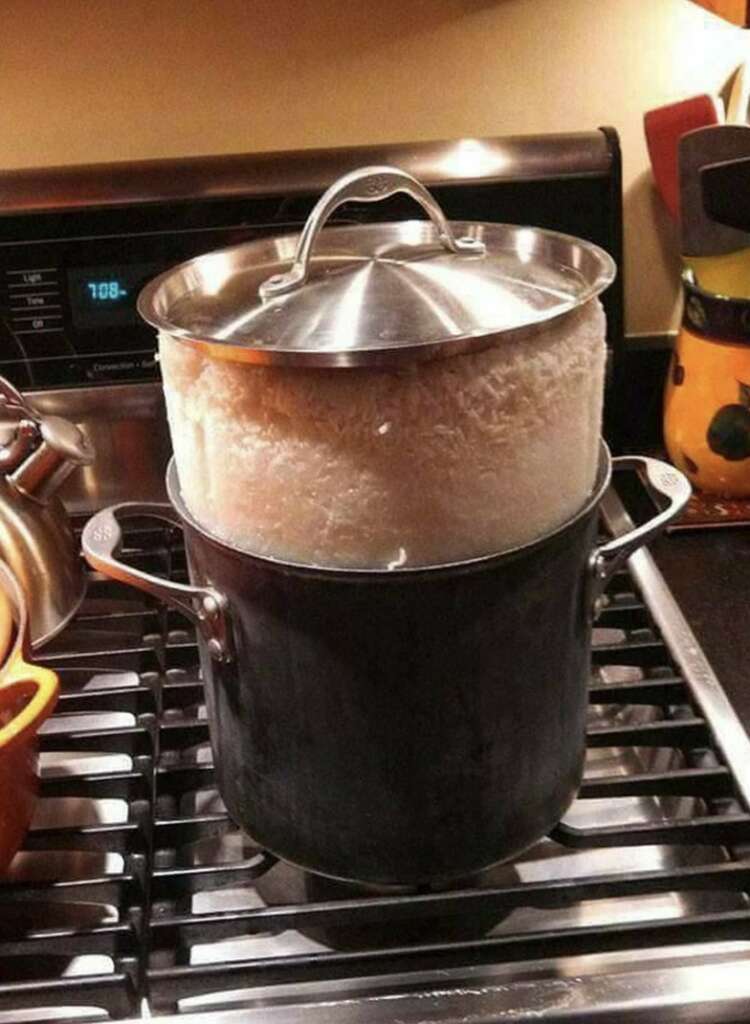 A black pot on a stove is overflowing with cooked rice, reaching the bottom of the lid. The lid is slightly raised due to the excess rice.