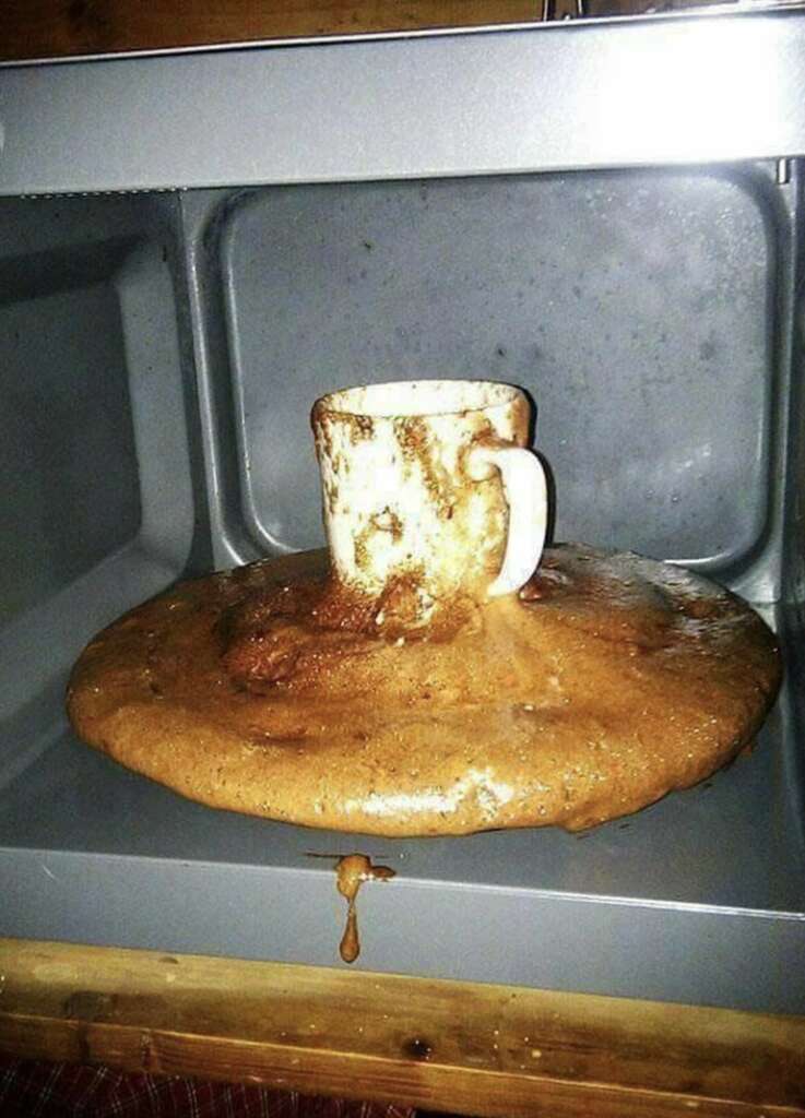 A chocolate cake has overflowed from a mug inside a microwave, spilling over onto the microwave's turntable and interior. The mug is partly coated with chocolate batter.