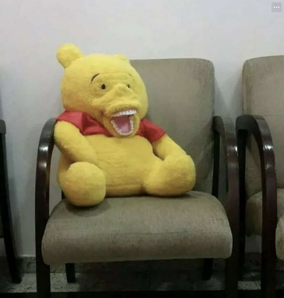 A plush toy resembling a yellow bear with a red shirt is sitting on a beige chair. The toy has an unusual wide-open mouth with visible teeth, creating a humorous and surprising expression. The chair is part of a set in a waiting area.