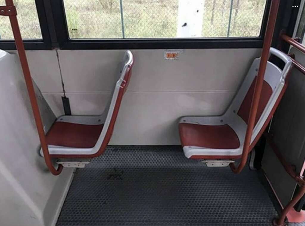 Two bus seats are positioned facing each other, separated by a narrow aisle. The seats have red cushions and are supported by metal frames. The bus has a gray floor with a textured surface, and a window is visible in the background.