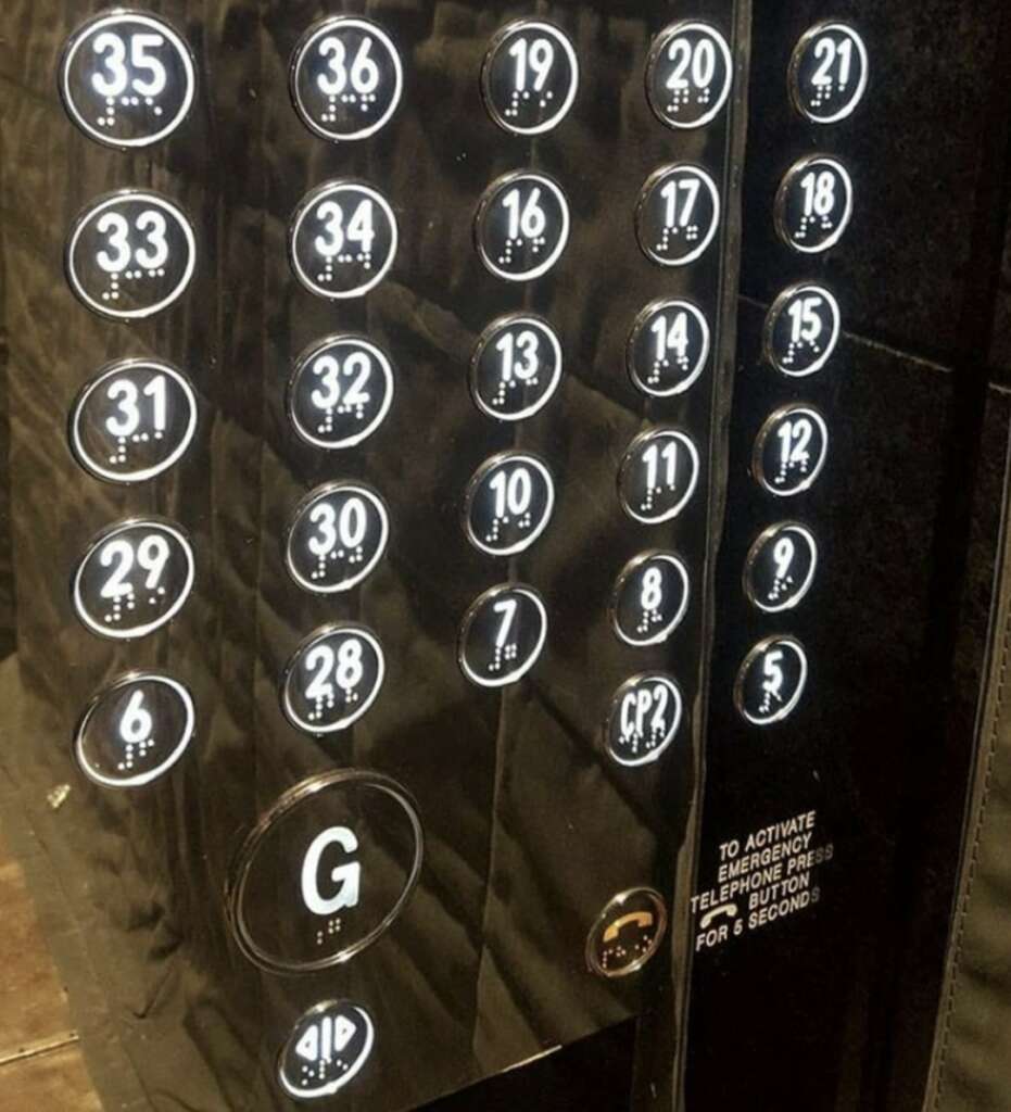 A close-up of an elevator panel with floors numbered from G to 36. The buttons are arranged in three columns with illuminated numbers. There's a note for activating emergency phone by pressing the button for 5 seconds.