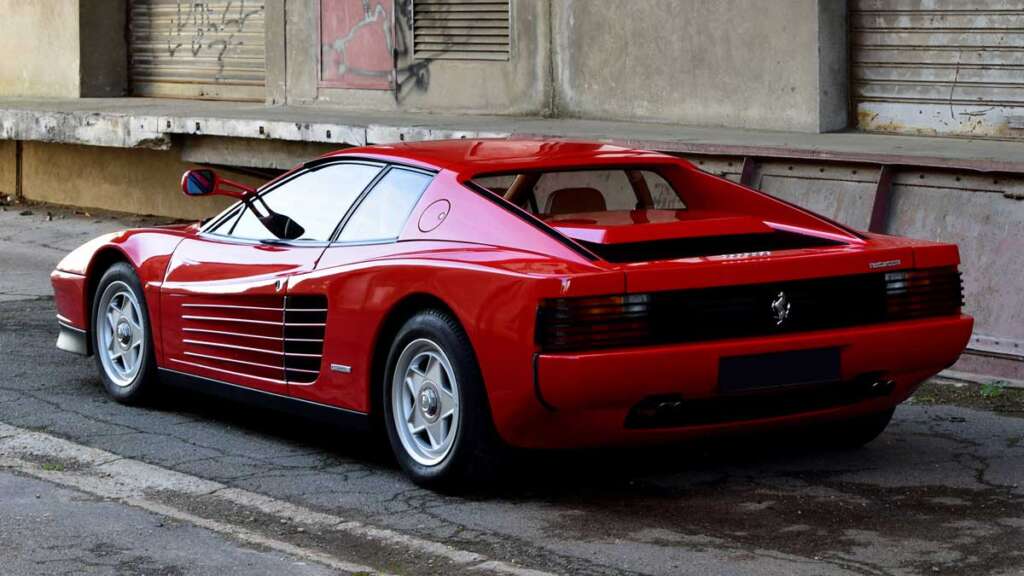 A red sports car, reminiscent of the coolest cars from the 1980s, is parked on an urban street. It boasts sleek, aerodynamic lines and a distinctive rear design. Industrial buildings with graffiti-splashed walls serve as its edgy backdrop.