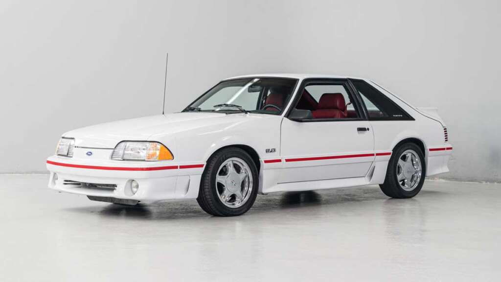 A white classic Ford Mustang with a red stripe and red interior is parked in a bright, minimalist indoor space. With its distinctive headlights and sleek body, this iconic car rivals the coolest cars from the 1980s, capturing vintage style and performance on silver wheels.