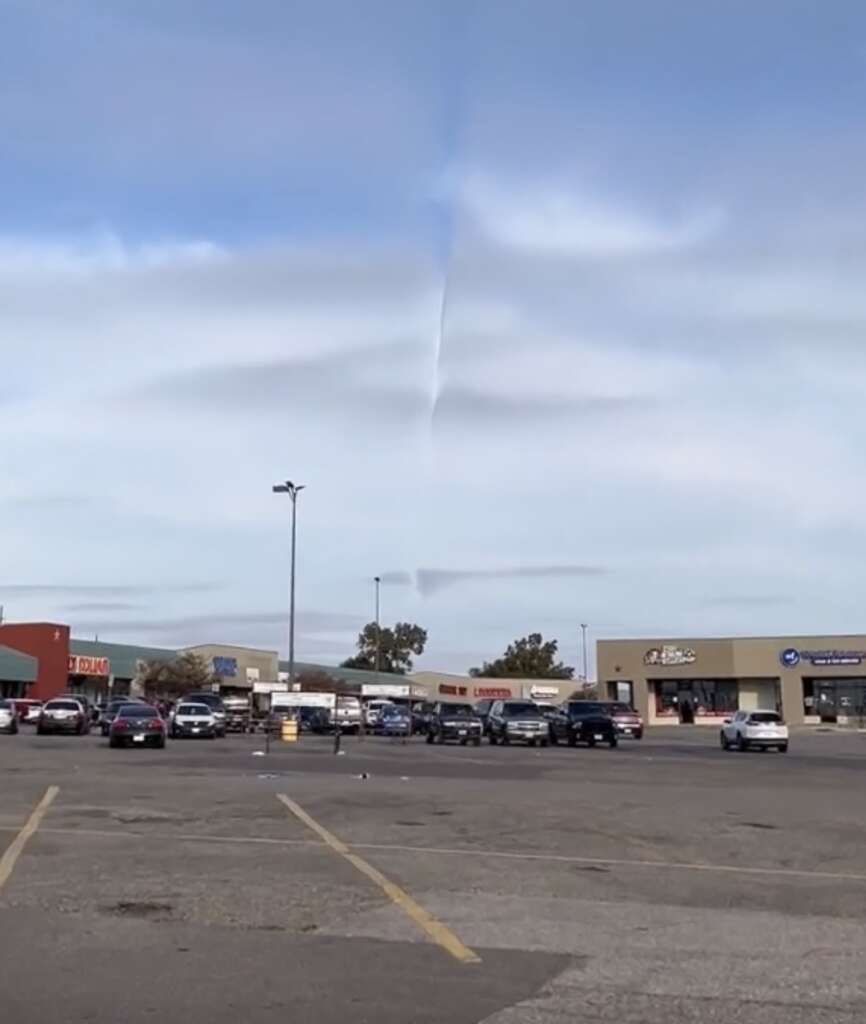 A strip mall parking lot with several parked cars under a cloudy sky. The distinctive clouds form a symmetrical, vertical line across the sky, creating a unique visual effect. Several storefronts are visible in the background.