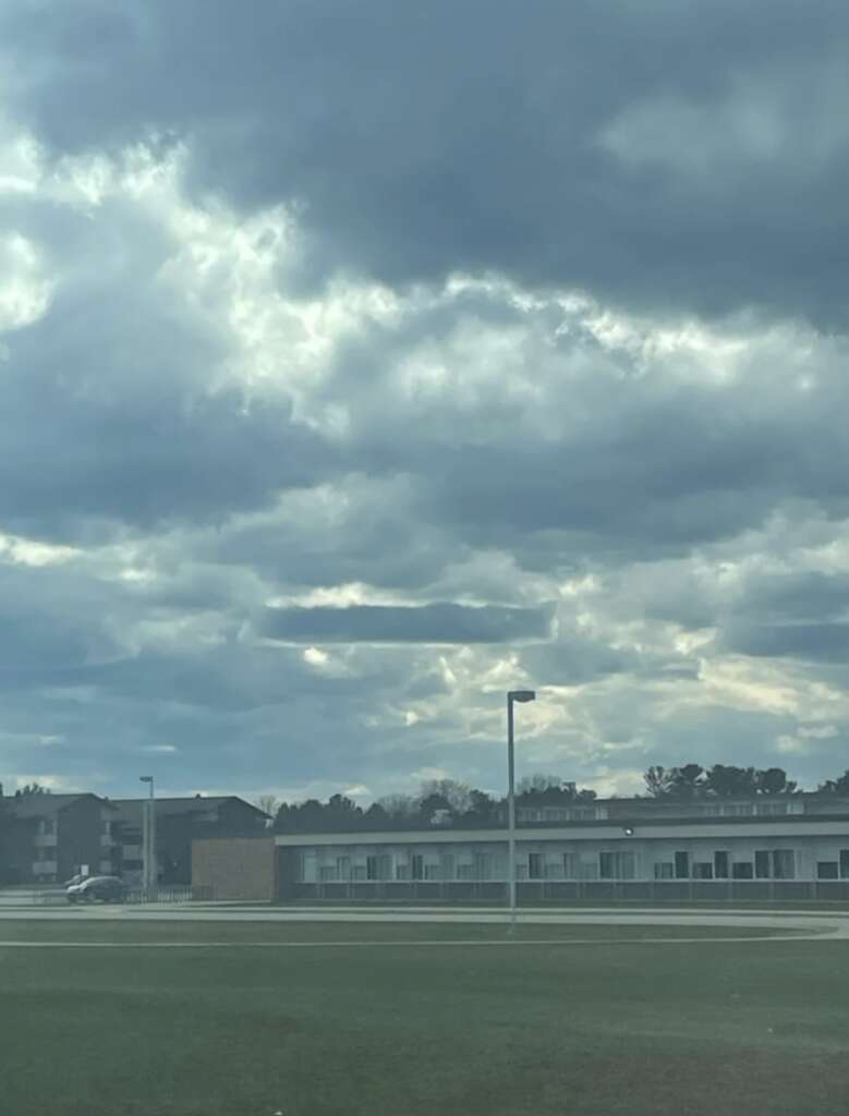 A cloudy sky with numerous fluffy gray clouds above a row of buildings in the distance. The foreground features a grassy field and streetlights, creating a calm, overcast scene.