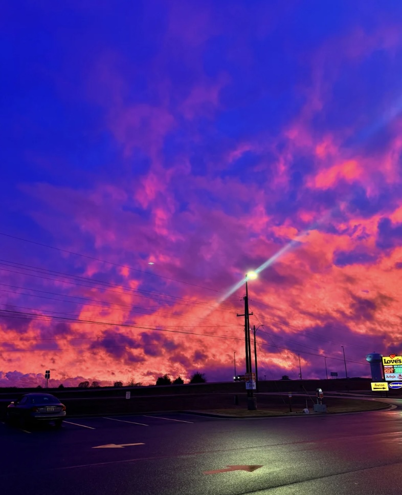 Vibrant sunset with pink and purple clouds fills the sky above a parking lot. A streetlight casts a glow on the wet pavement, and a few cars are visible in the distance. A brightly lit sign can be seen on the right side.