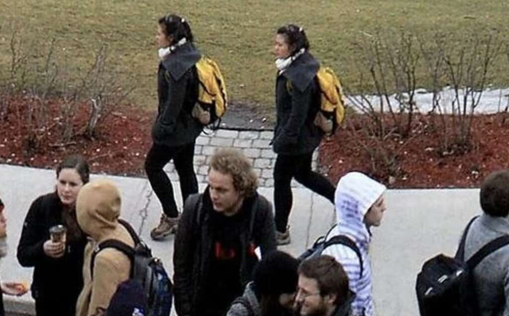 A group of people, mostly wearing backpacks and casual winter clothing, walk along a path in a park. Two individuals with yellow backpacks are walking side by side, while others engage in conversation. The ground is partially covered with mulch and grass.